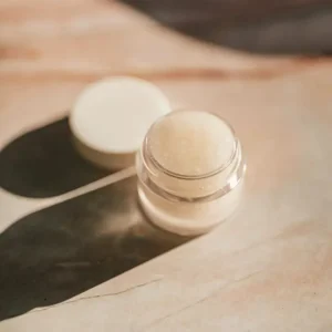 Small frosted glass pot of balm with its white lid beside it, in hard sunlight on a warm beige surface